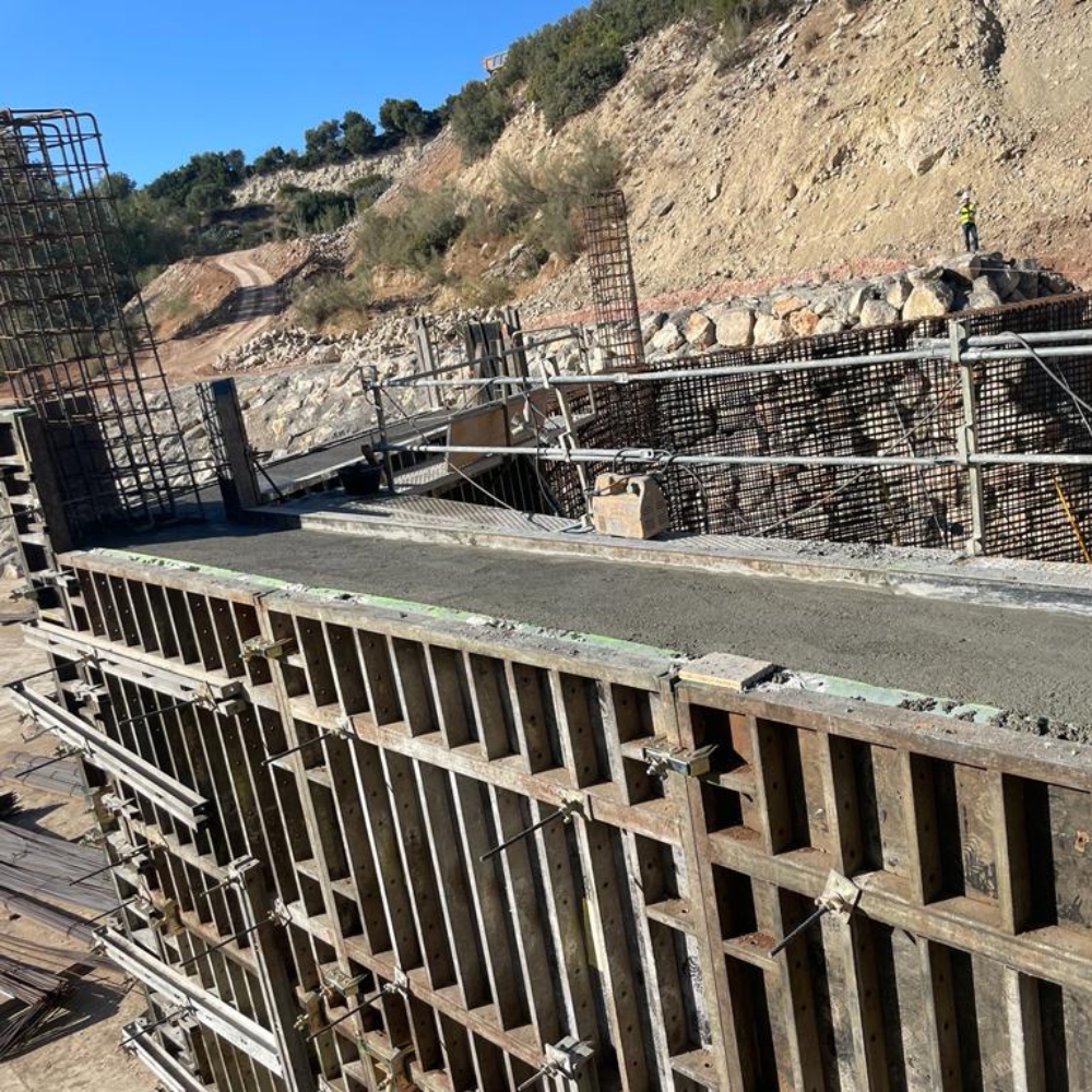 Proceso de Encofrado en Obra de Construcción magen que muestra el proceso de encofrado en una obra de construcción, donde se observa el moldeado de las estructuras de hormigón. Las planchas de encofrado están dispuestas para contener el vertido de hormigón en áreas específicas, asegurando la formación precisa de las paredes y columnas. Al fondo, se aprecian las formaciones rocosas y terrenos irregulares que rodean el sitio de la obra, destacando la adaptación de la construcción al entorno.