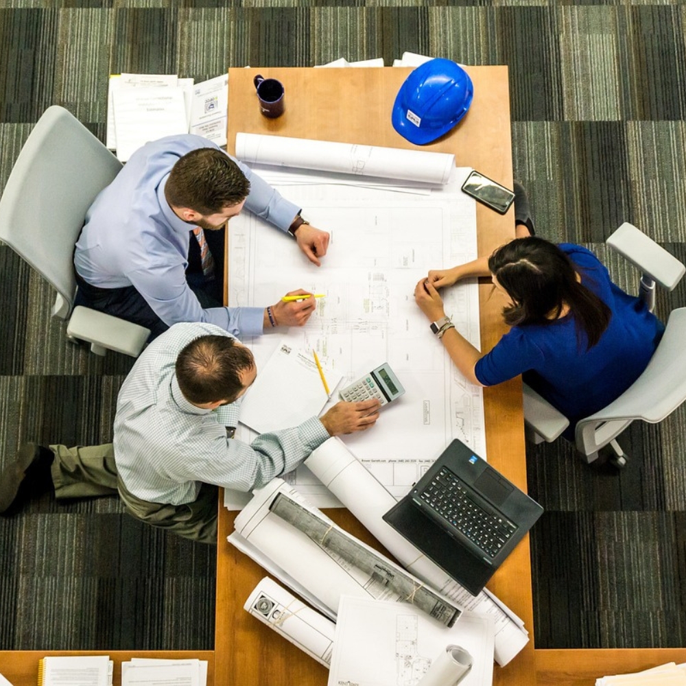 Reunión de Planificación de Proyecto de Construcción Vista aérea de un equipo de profesionales revisando planos en una mesa de trabajo. Los participantes discuten detalles técnicos y de planificación del proyecto, apoyados por herramientas como calculadoras, ordenadores y planos arquitectónicos. La reunión refleja la colaboración y el análisis riguroso necesarios para llevar a cabo un proyecto de construcción exitoso.