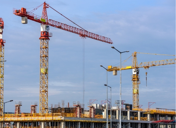 Varias grúas torre operando en un gran proyecto de construcción de edificios de varios pisos. Las estructuras de acero y concreto están en una fase avanzada de desarrollo, con andamios y maquinaria pesada en el sitio. El cielo despejado y las grúas en movimiento reflejan el ritmo acelerado del crecimiento urbano.