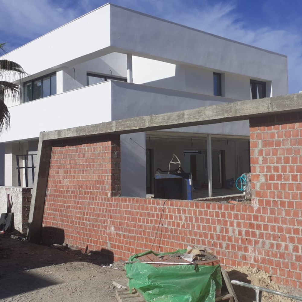 Construcción de Viviendas Unifamiliares Pareadas en El Altillo, Jerez Vista de una obra en proceso de construcción de viviendas unifamiliares pareadas en El Altillo, Jerez. La estructura moderna de dos pisos presenta líneas limpias y minimalistas, mientras los muros exteriores de ladrillo aún están en desarrollo. El proyecto avanza hacia su finalización, con los acabados exteriores comenzando a tomar forma bajo un cielo despejado.