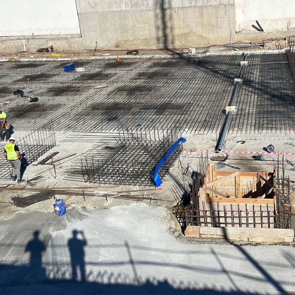 Proceso de Ferrallado en Proyecto de Construcción Vista de la fase de ferrallado en una obra de construcción, donde se están instalando y organizando las barras de acero para refuerzo de la cimentación. Los trabajadores revisan la disposición de los enrejados de acero, que servirán como soporte estructural para el hormigón. La imagen captura un momento clave en la preparación de la base de la edificación, esencial para garantizar la estabilidad y durabilidad de la construcción.