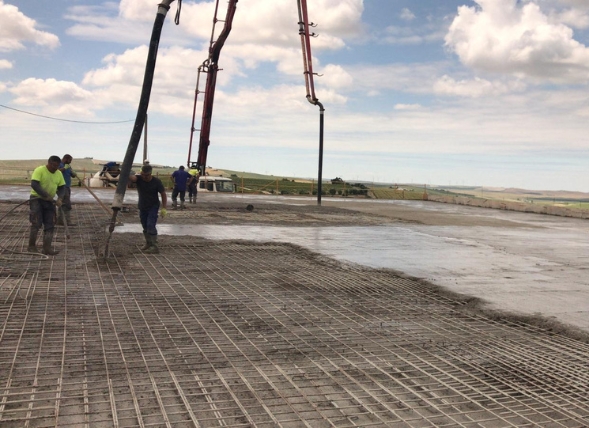 Trabajadores de la construcción supervisando el proceso de colado de hormigón en una estructura de encofrado. Un gran cubo de hormigón suspendido facilita la descarga del material, mientras los obreros ajustan los detalles en la fase de cimentación. El sitio está organizado con andamios y equipos especializados para garantizar la seguridad y eficiencia en el desarrollo de la obra.