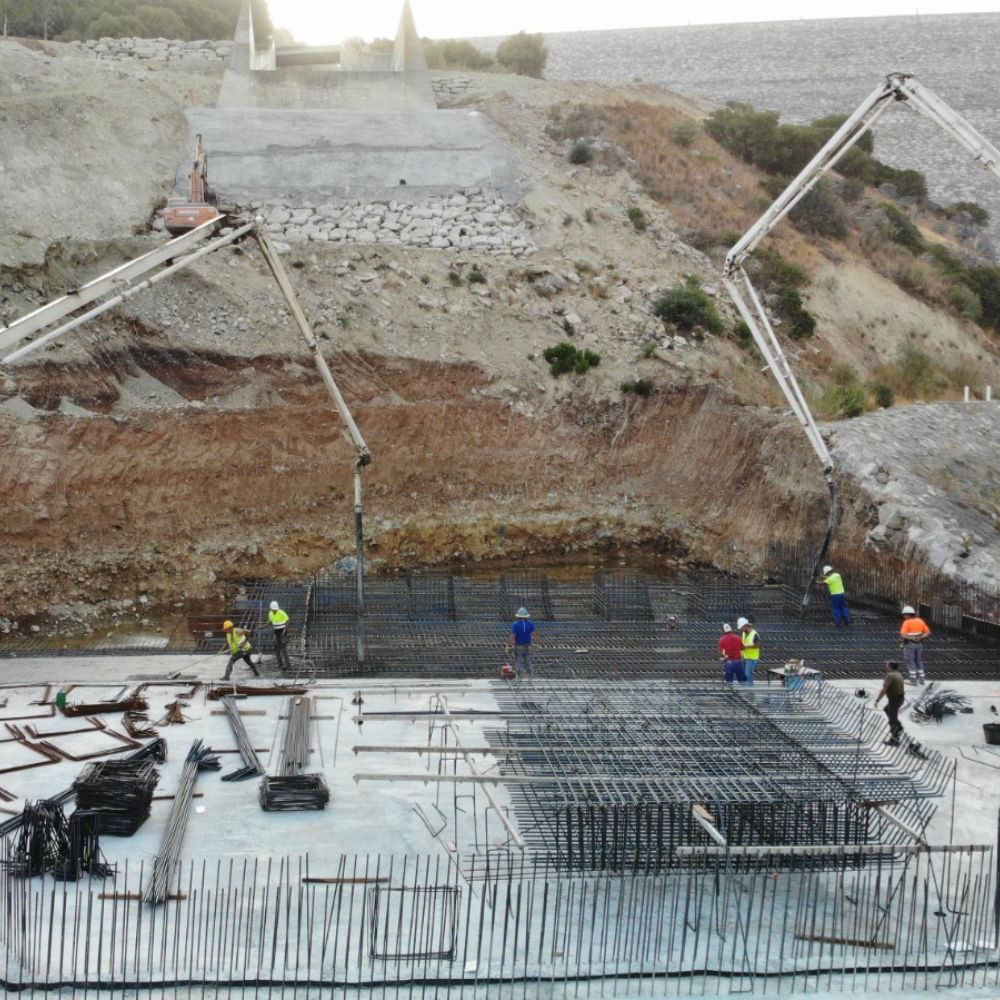 Imagen del colado de concreto en una sección de la presa de Guadalcacín. Se observan camiones bomba vertiendo concreto sobre una estructura de acero de refuerzo, con varios trabajadores en el sitio supervisando y ajustando la distribución del material. Al fondo, una pendiente excavada y revestida de roca completa la escena de construcción.