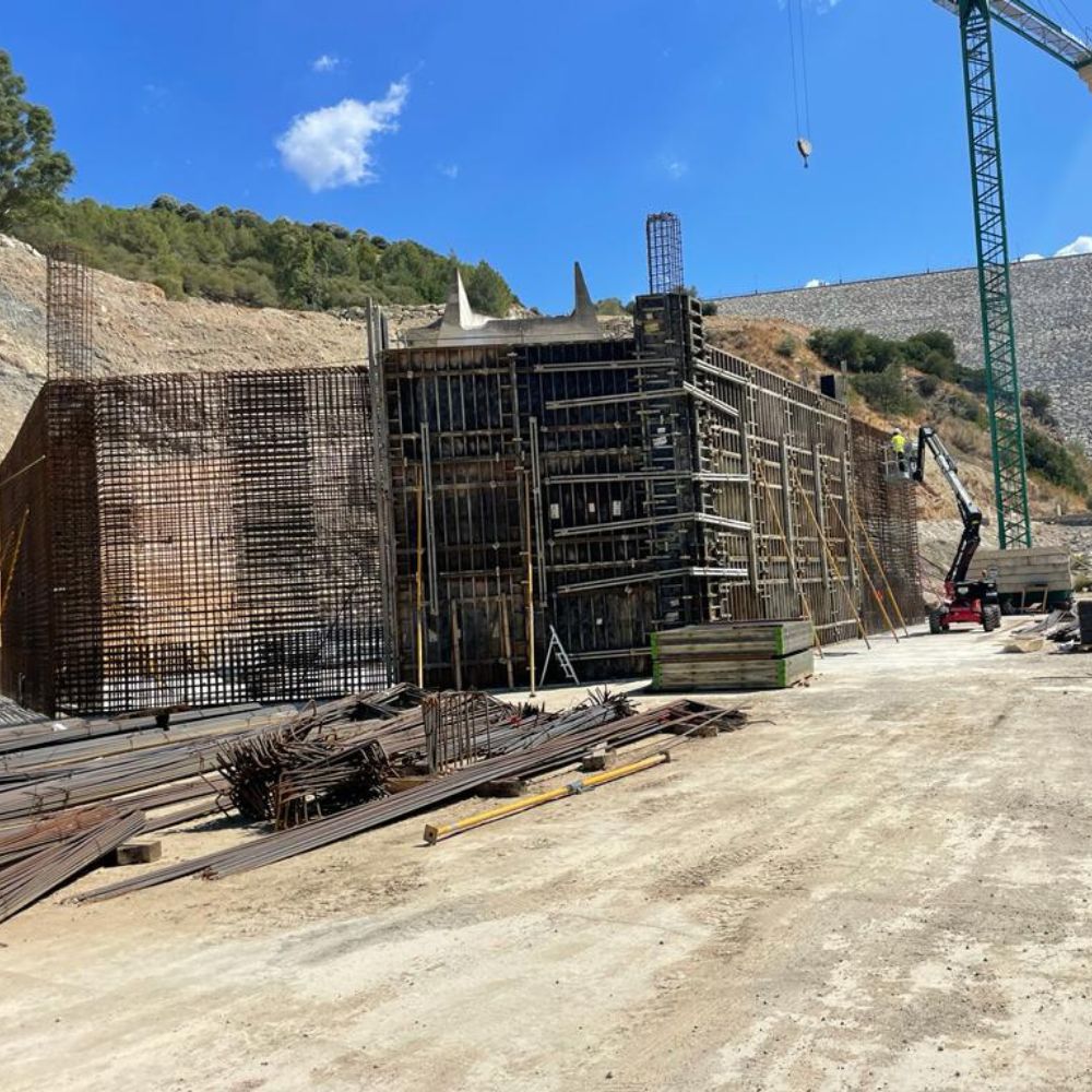 Imagen del proceso de construcción en la presa de Guadalcacín. Se observa una estructura de refuerzo de acero enmarcada por un encofrado de madera, rodeada de materiales de construcción y herramientas. Una grúa y un elevador están en operación mientras los trabajadores ensamblan y ajustan los elementos estructurales en el lugar.